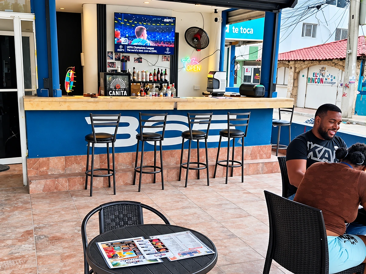 The 365 Beach Bar in daylight — the blue bar front lettered 365, timber counter, bar stools, a wall-mounted TV showing live football and the neon OPEN sign, with guests seated at a patio table