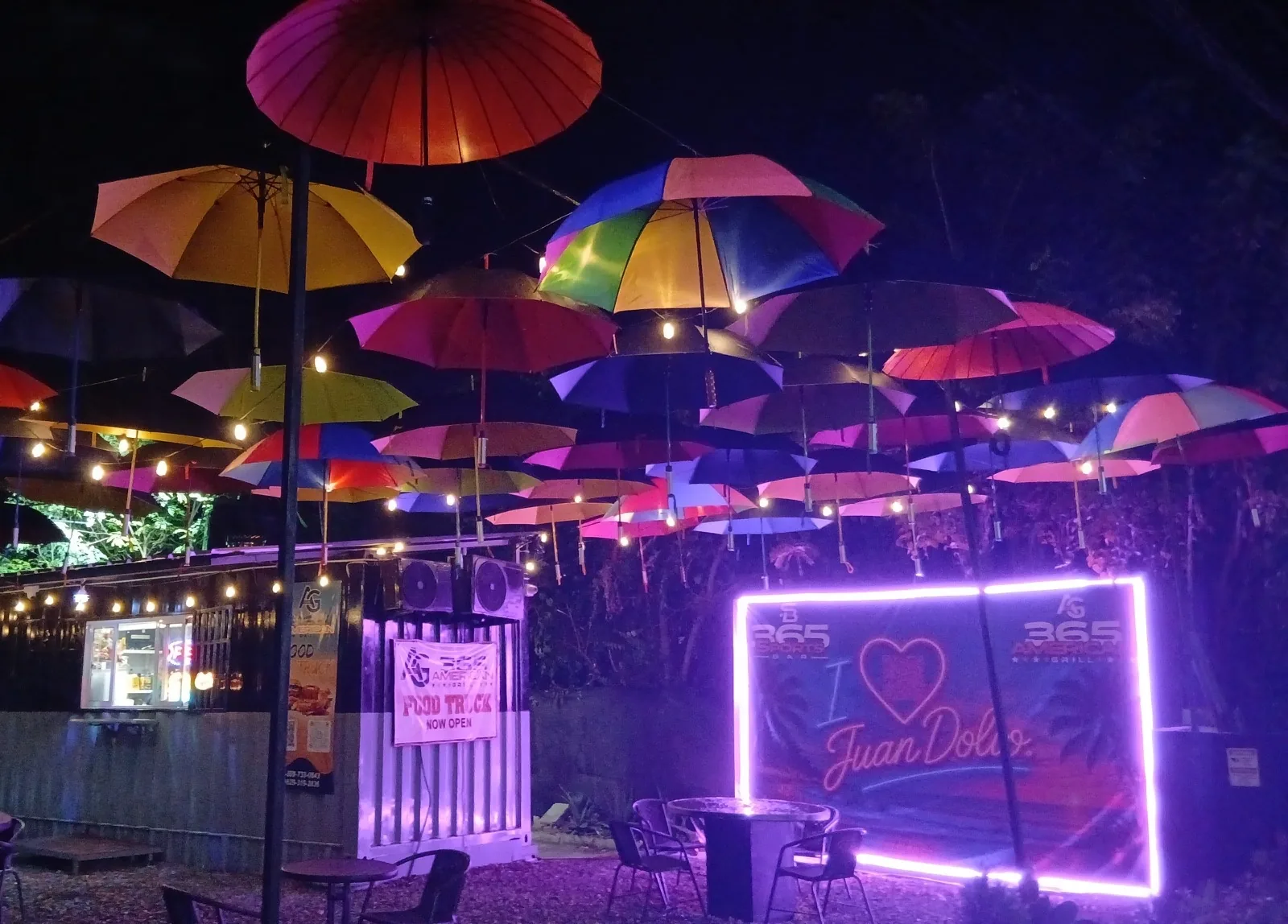 The 365 American Grill umbrella patio at night — colourful umbrellas and string lights over the food truck window and the neon I love Juan Dolio sign